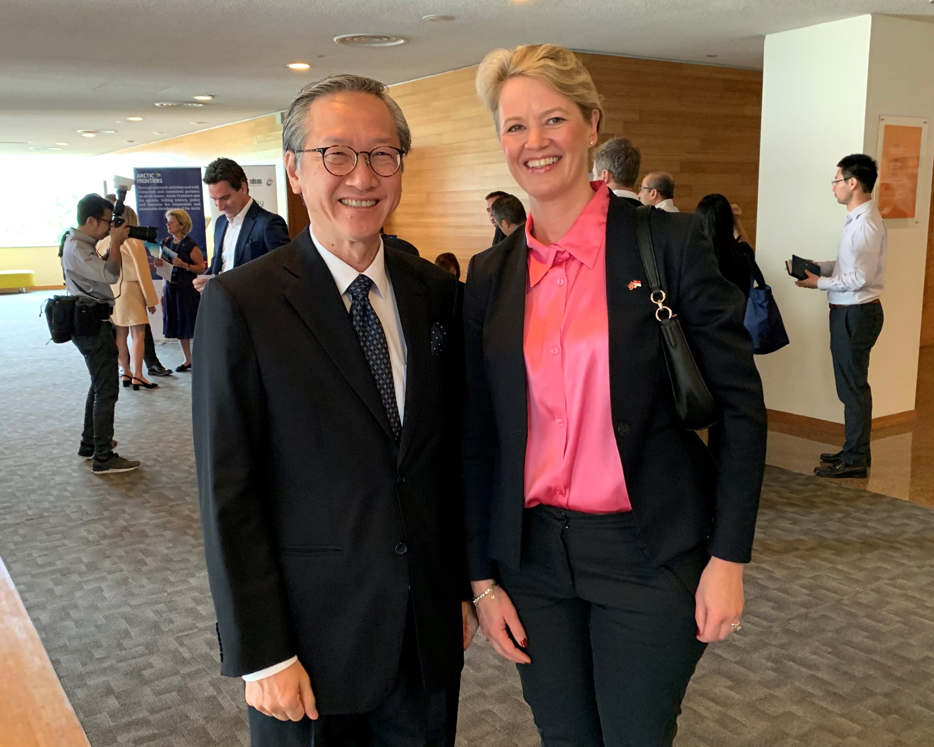 Two people in business attire standing indoors; others visible in background.